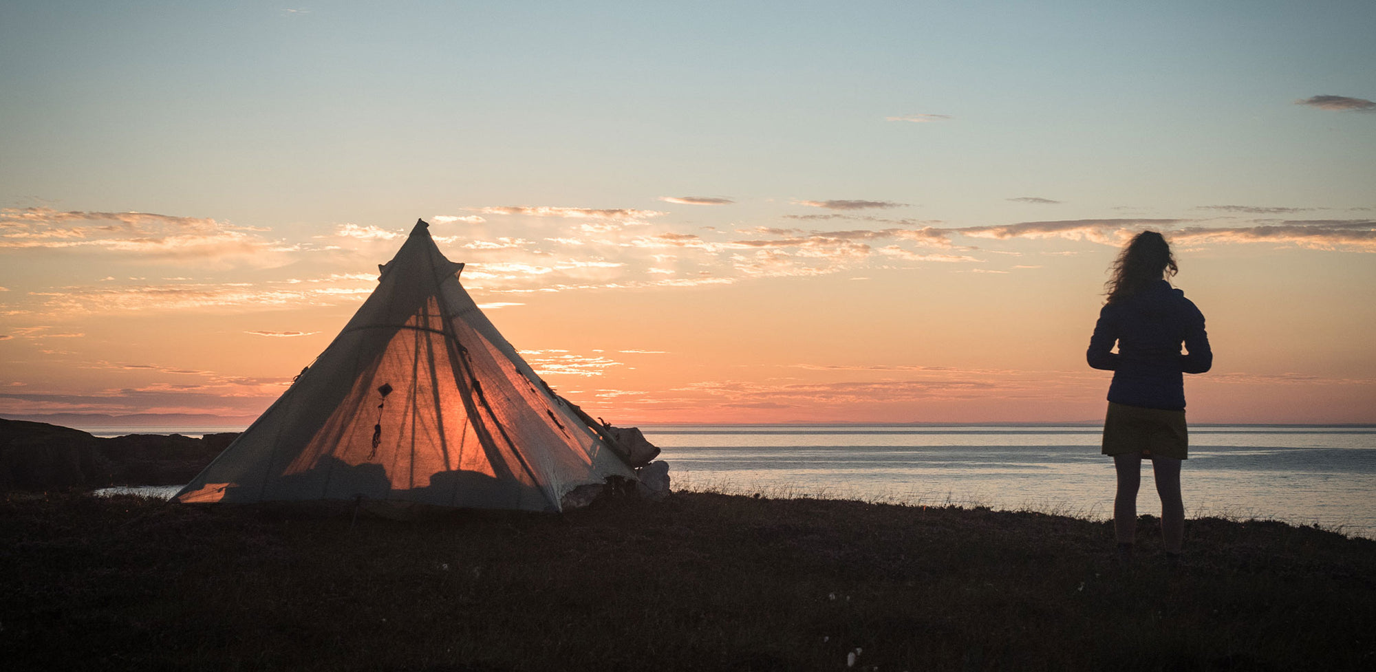 A person standing in front of a teepee at sunset.