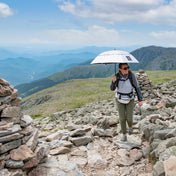 A hiker traverses rocky terrain with the Hyperlite Mountain Gear Runabout Bundle in tow, wearing the Versa around their waist and using the umbrella