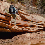 Hiker skirts the edge of a sandstone ledge above some water wearing the Hyperlite Mountain Gear Unbound 55