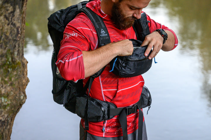A bearded man adjusting his backpack near a body of water.