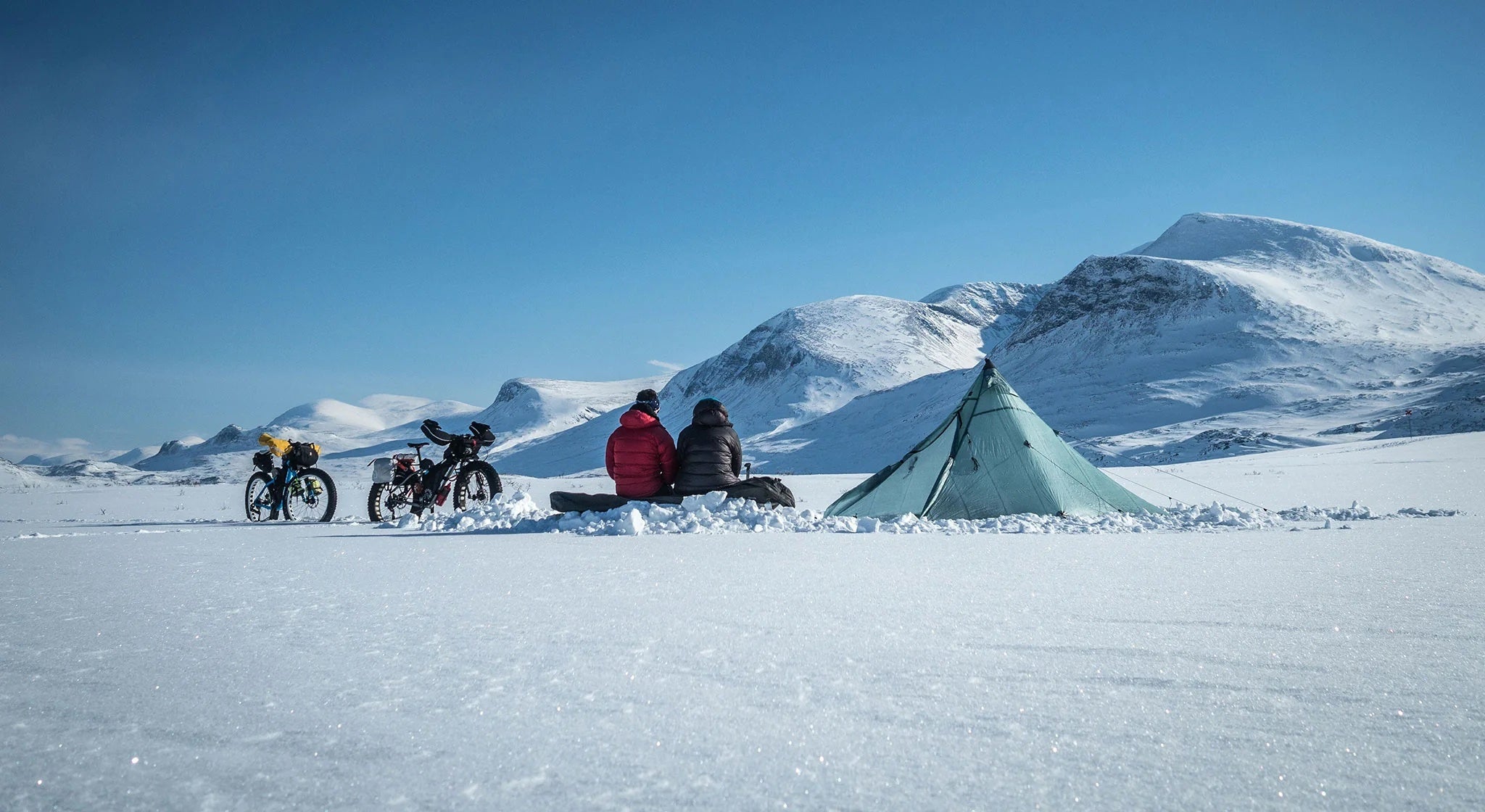 Three people sitting beside a tent on a snowy landscape with bicycles nearby, with mountains in the background.