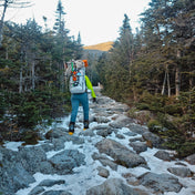 A hiker ascends a snowy and rocky hill in between the trees using Hyperlite Mountain Gear