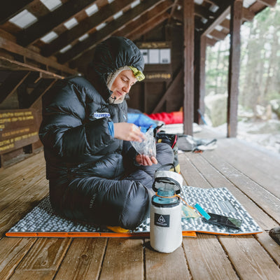 A camper sits in a lean to with the Hyperlite Mountain Gear Insulator during winter time