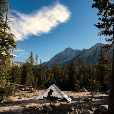 A hiker relaxes at camp amongst the trees in the Hyperlite Mountain Gear Mid 1 Tarp