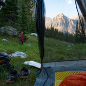 View from the interior of Hyperlite Mountain Gear Shelters UltaMid 2 Half Insert without tent covering overlooking a grassy mountain setting. A pair of hiking shoes sits in the foregrounds, and a model stands in the background
