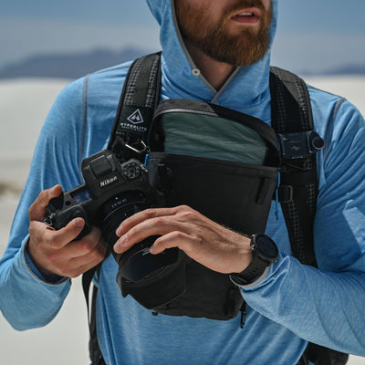 A hiker with the Hyperlite Mountain Gear Camera Pod attached to the pack at the chest with a camera in hand